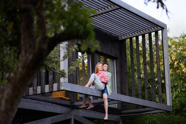 A young couple sitting and cuddling in hammock terrace in their new home in tiny house in woods, sustainable living concept. - HPIF05025