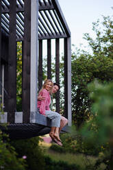 Ein junges Paar sitzt und kuschelt in der Hängematte auf der Terrasse ihres neuen Hauses in einem winzigen Haus im Wald, ein nachhaltiges Wohnkonzept. - HPIF05026