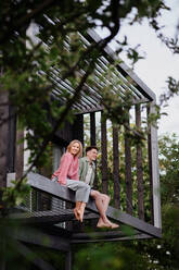 A young couple sitting and cuddling in hammock terrace in their new home in tiny house in woods, sustainable living concept. - HPIF05027