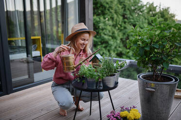 A young woman taking care of her herbs, watering them on terrace in tiny house, sustainable living. - HPIF05036