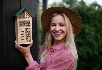 A young woman garedener holding bug and insect hotel on terrace in garden, sustainable lifestyle. - HPIF05044