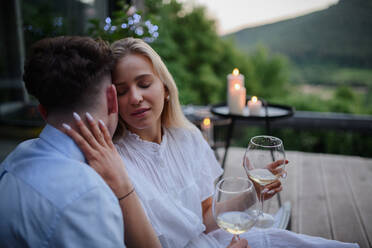 A young couple with wine resting outdoors on terrace in evening, weekend away in tiny house in countryside, sustainable living. - HPIF05055