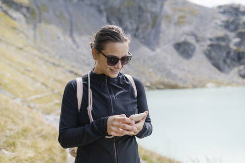 Lächelnder Wanderer mit Mobiltelefon in der Nähe eines Bergsees - MEF00146