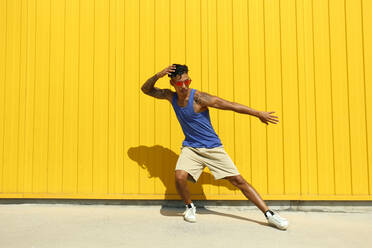 Junger Mann mit Sonnenbrille tanzt auf dem Fußweg vor einer gelben Wand - SYEF00043