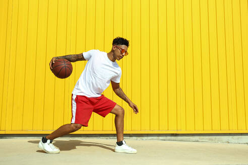 Young man playing with basketball on footpath in front of yellow wall - SYEF00055