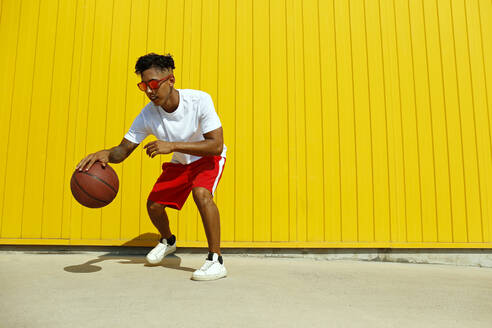 Mann mit roter Sonnenbrille spielt mit einem Basketball auf dem Fußweg vor einer gelben Wand - SYEF00056