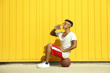 Man drinking water from bottle sitting on basketball in front of yellow wall - SYEF00062