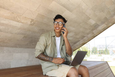 Smiling freelancer talking through mobile phone sitting with laptop on bench - SYEF00067