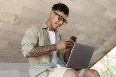 Happy freelancer using smart phone sitting with laptop - SYEF00070