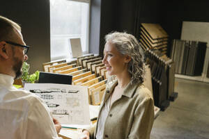 Lächelnde Geschäftsfrau betrachtet einen Kollegen, der einen Bauplan in einem Architekturbüro hält - SEAF01526