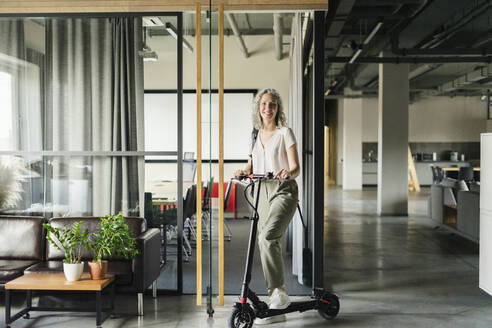 Portrait of smiling businesswoman with electric scooter in office - SEAF01619