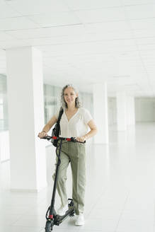 Portrait of smiling businesswoman with electric scooter in office - SEAF01620