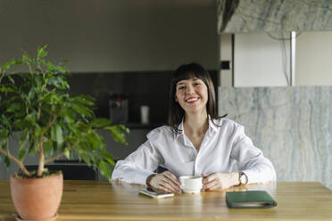 Portrait of smiling businesswoman sitting at table in office kitchen - SEAF01625