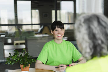 Smiling woman in green t-shirt looking at colleague in office - SEAF01670