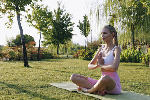 Lächelnde Frau übt Padmasana-Pose auf Übungsmatte im Park - SYEF00091