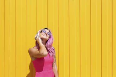 Glückliche junge Frau, die über drahtlose Kopfhörer vor einer gelben Wand Musik hört - SYEF00103
