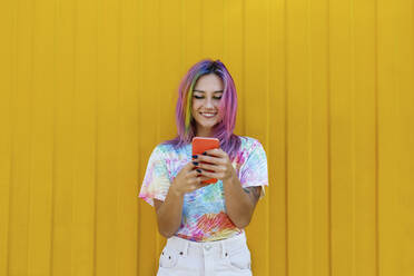 Smiling young woman wearing tie dye t-shirt using smart phone in front of yellow wall - SYEF00105