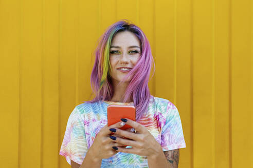 Lächelnde junge Frau mit Mobiltelefon vor gelber Wand - SYEF00107