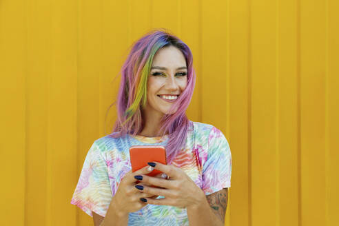 Glückliche junge Frau, die ihr Handy vor einer gelben Wand hält - SYEF00109