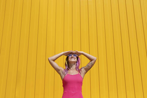 Glückliche Frau in rosa Kleid mit Augenschutz vor einer gelben Wand - SYEF00111