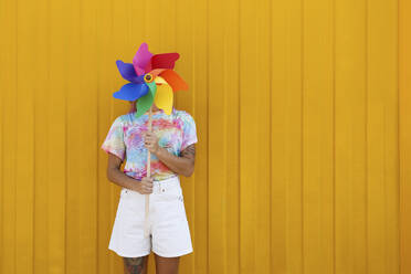 Young woman covering face with rainbow colored pinwheel toy in front of yellow wall - SYEF00121