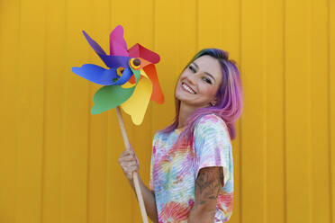 Happy young woman holding multi colored pinwheel toy in front of yellow wall - SYEF00125