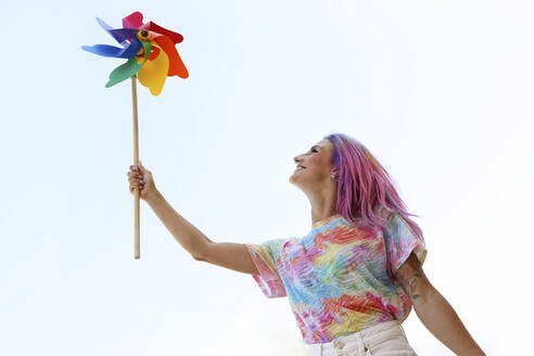 Glückliche junge Frau hält regenbogenfarbenes Windradspielzeug unter klarem Himmel - SYEF00129
