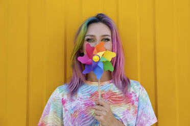 Woman covering mouth with colorful pinwheel toy in front of yellow wall - SYEF00131