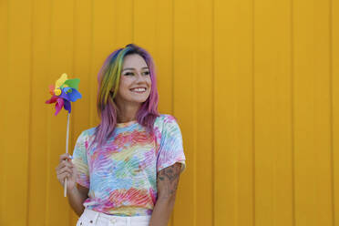 Happy woman holding colorful pinwheel toy in front of yellow wall - SYEF00134