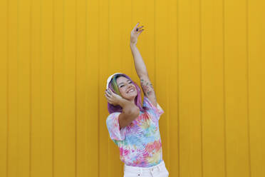 Happy woman enjoying music listening through wireless headphones and dancing in front of yellow wall - SYEF00136