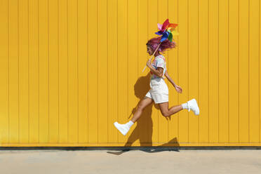 Young woman holding colorful pinwheel toy jumping on footpath in front of yellow wall - SYEF00141