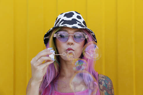 Young woman wearing hat blowing soap bubbles in front of yellow wall - SYEF00144