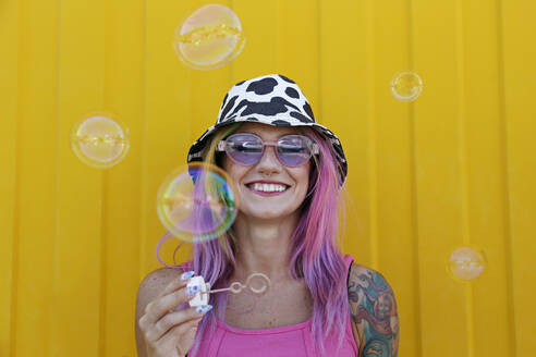 Happy woman holding bubble wand in front of yellow wall - SYEF00145