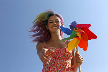 Glückliche Frau hält buntes Windrad-Spielzeug und gestikuliert unter blauem Himmel - SYEF00147