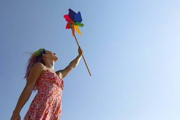 Glückliche Frau mit erhobener Hand, die ein buntes Windrad-Spielzeug hält, gestikuliert unter blauem Himmel - SYEF00148