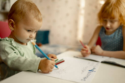 Cute baby girl drawing on paper with sister doing homework at table - ANAF00695
