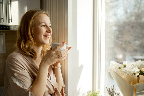 Glückliche junge Frau mit Kaffeetasse, die durch ein Fenster hinausschaut - ANAF00697