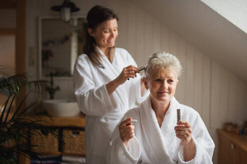 Eine glückliche ältere Mutter im Bademantel mit erwachsener Tochter, die zu Hause Lockenwickler benutzt, Selbstpflegekonzept. - HPIF05160