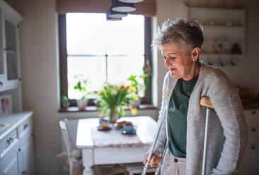 A senior woman with crutches indoors at home, walking in kitchen. - HPIF05179