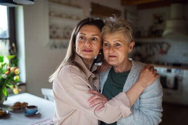Ein Porträt einer älteren Mutter, die sich mit ihrer erwachsenen Tochter zu Hause umarmt und zur Seite schaut. - HPIF05183