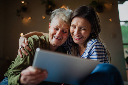 Ein Porträt einer glücklichen älteren Mutter mit ihrer erwachsenen Tochter, die zu Hause ein Tablet benutzt. - HPIF05200