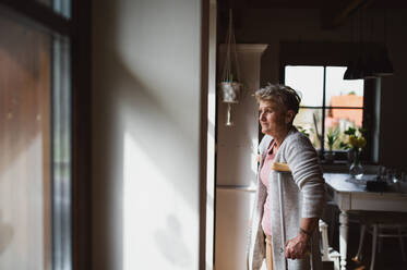 A sad senior woman with crutches indoors at home, looking out through window. - HPIF05212