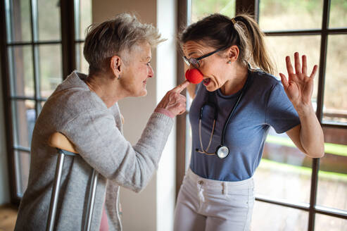 Eine Pflegekraft oder ein Pfleger mit roter Nase besucht eine ältere Frau zu Hause und hat Spaß. - HPIF05226