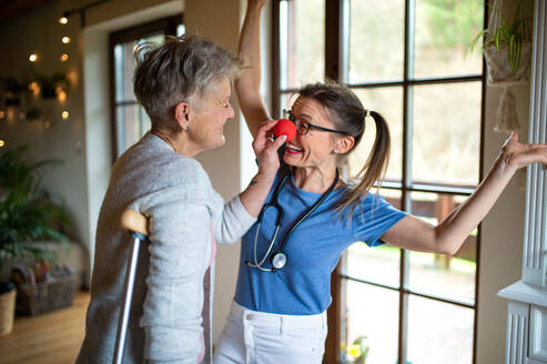 Eine Pflegekraft oder ein Pfleger mit roter Nase besucht eine ältere Frau zu Hause und hat Spaß. - HPIF05227