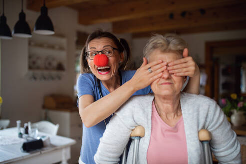 Gesundheits- und Krankenpfleger mit roter Nase, der eine ältere Frau zu Hause besucht, sich amüsiert und ihr die Augen zuhält. - HPIF05228