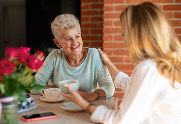 Eine glückliche ältere Mutter unterhält sich mit ihrer erwachsenen Tochter, während sie zu Hause sitzt und Kaffee trinkt. - HPIF05240