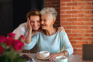 Eine glückliche ältere Mutter, die sich mit ihrer erwachsenen Tochter umarmt, während sie zu Hause sitzt und Kaffee trinkt. - HPIF05241