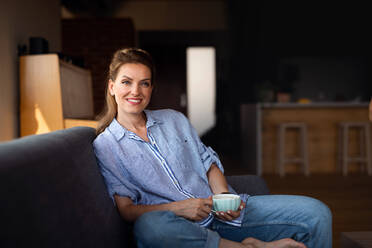 Ein Porträt der glücklichen Mitte erwachsene Frau mit Kaffee lächelnd und sitzen auf Sofa drinnen zu Hause - HPIF05250
