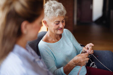 A happy senior woman sitting on sofa and learning to knit her adult daughter indoors at home. - HPIF05251