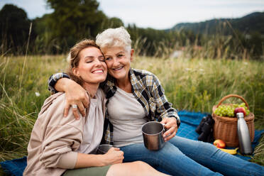 Eine glückliche ältere Mutter, die ihre erwachsene Tochter umarmt, während sie draußen in der Natur sitzt und ein Picknick macht. - HPIF05276